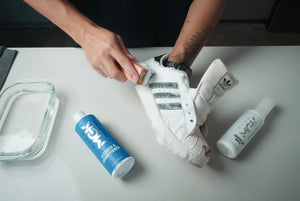 White shoe with black stripes has soap lather on the side with a bottle of Shoe MGK shoe cleaner and Shoe MGK white touch up. There is a glass container with water to the side of the bottle.